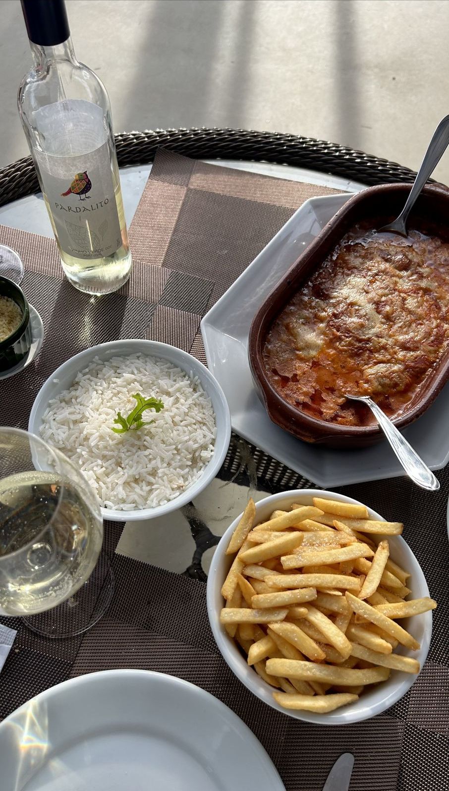 Una mesa con una botella de vino, un cuenco de arroz, un plato de papas fritas y un plato al horno.