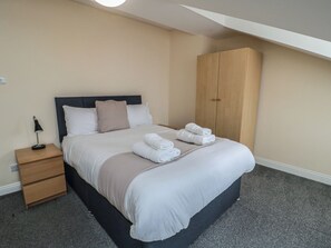 Ein ordentlich bezogenes Bett mit weißen Leinen und einer beigen Tagesdecke, ein Nachttisch aus Holz mit einer Lampe und ein Holzschrank in einem Hotelzimmer.