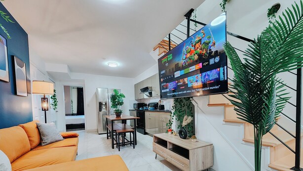 Una sala de estar moderna con un sofá, un televisor montado en la pared, una zona de cocina y una escalera.