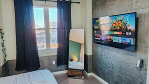 Una habitación de hotel con cama, televisor montado en la pared mostrando una selección de películas, ventana con cortinas y vistas a la ciudad.
