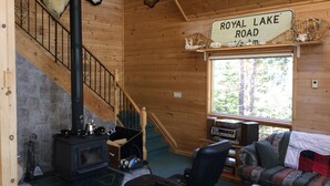 A log cabin interior with a fireplace, a sign reading "ROYAL LAKE ROAD", a staircase, a couch, and a window with a view of trees. - Royal Lake Cabin (Royal Lake)