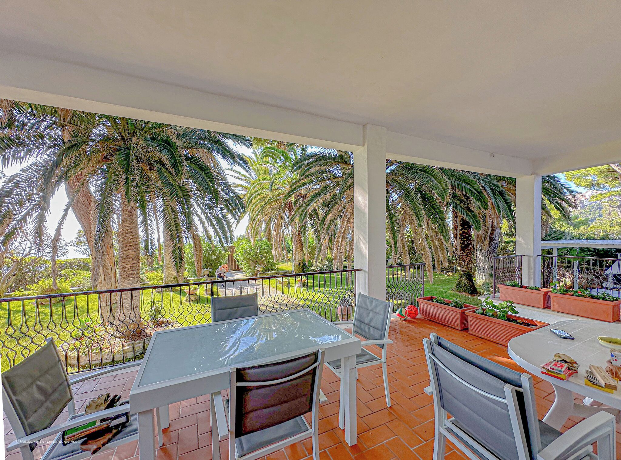 Une terrasse extérieure couverte, dotée d’une table et de chaises, entourée d’une clôture en métal et de palmiers.