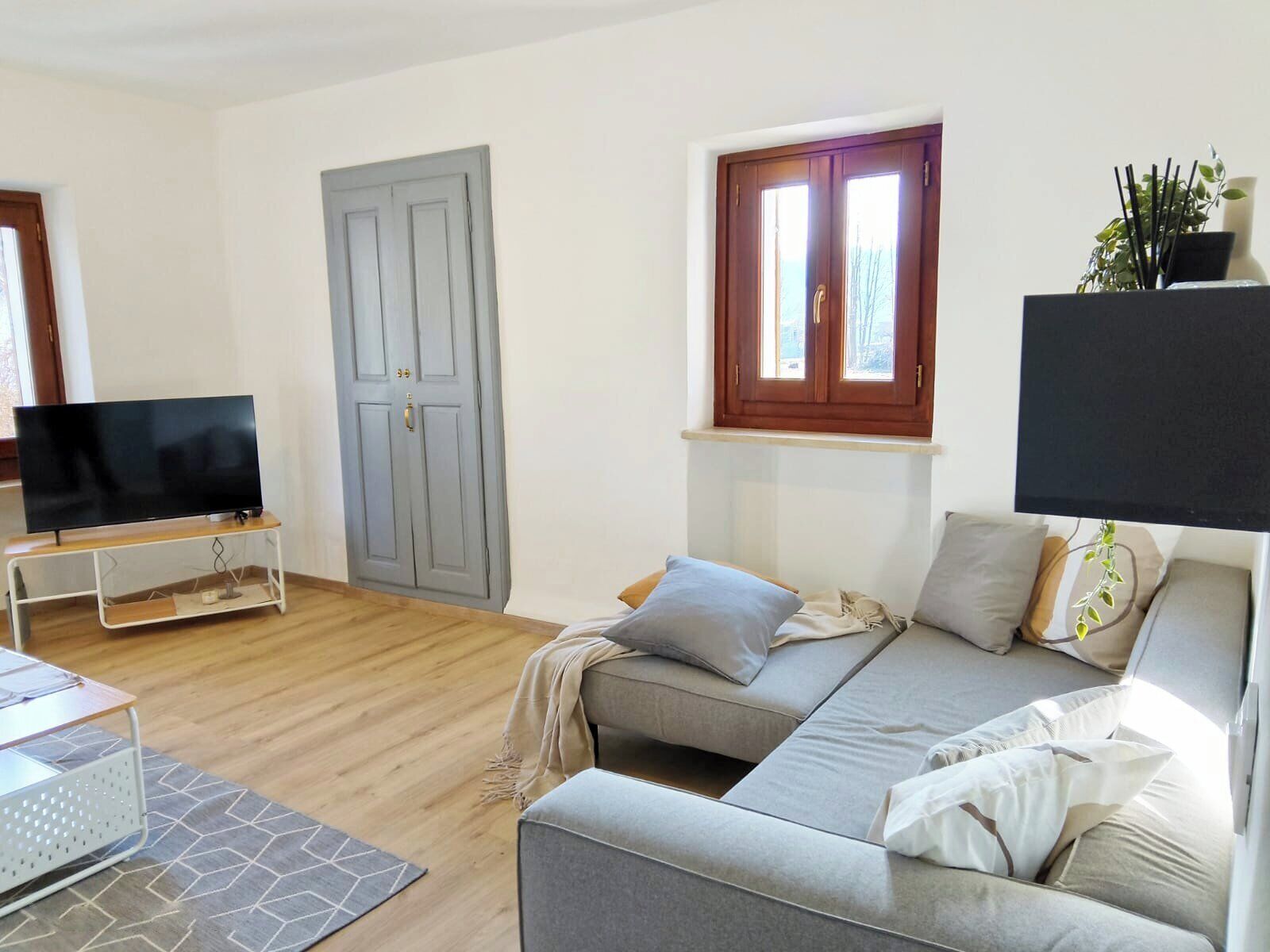 A living room with a grey sofa, a television on a stand, a wooden floor, a grey patterned rug, and a window with a wooden frame.