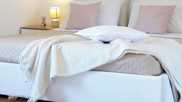 A neatly made bed with a white comforter and pillows, a bedside lamp, and a small table with a candle and a container. - QuickHome Appartamento Gelsomino (Andorno Micca)
