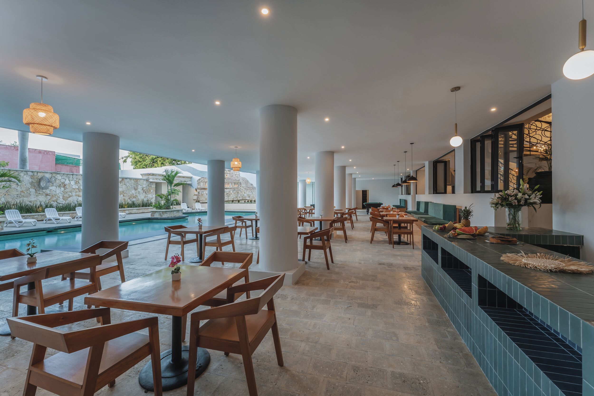 A restaurant with wooden tables and chairs, a pool in the background, and a counter with food items.