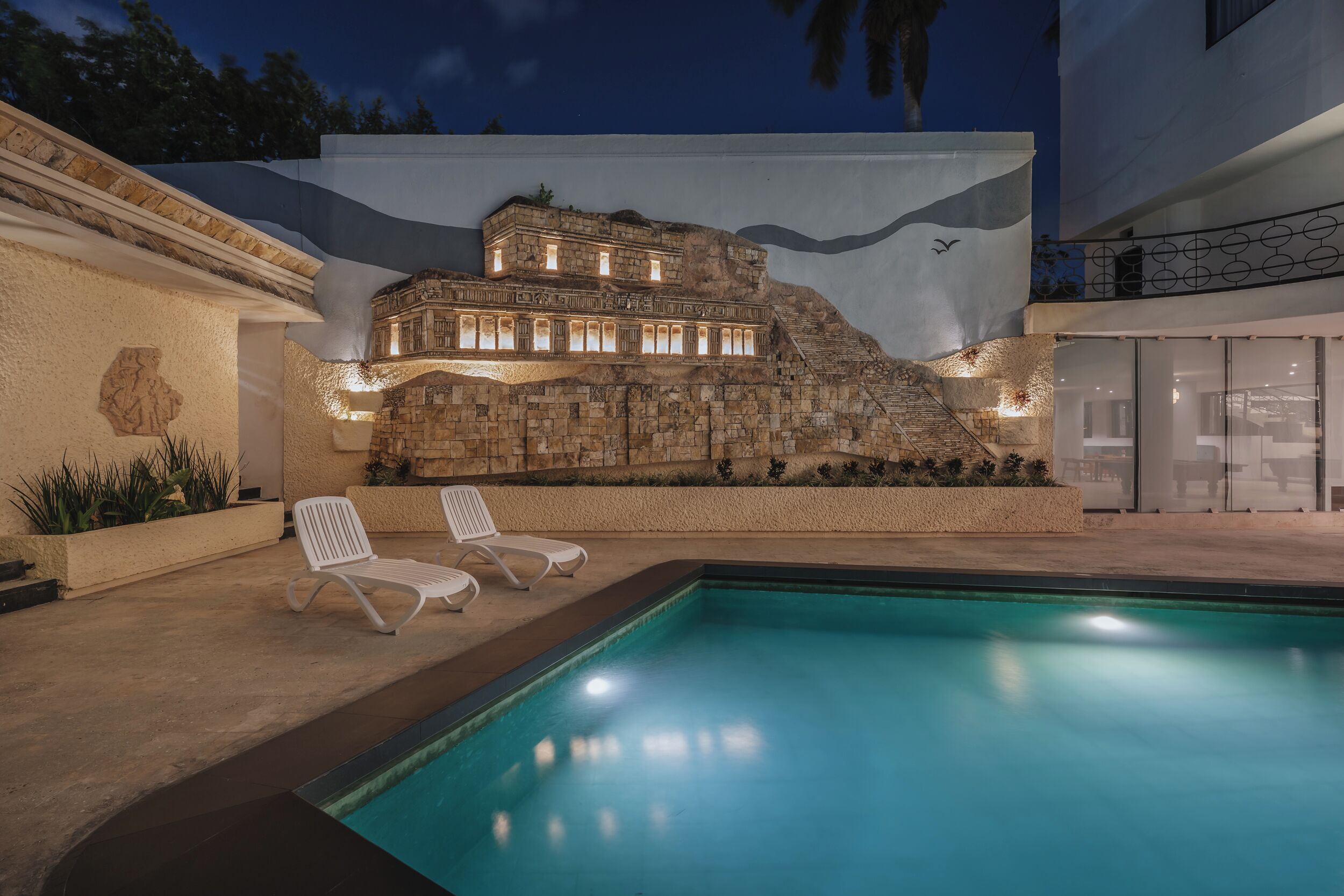 A swimming pool with two lounge chairs and a rock wall with a building on top.