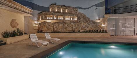 A swimming pool with two lounge chairs and a rock wall with a building on top.