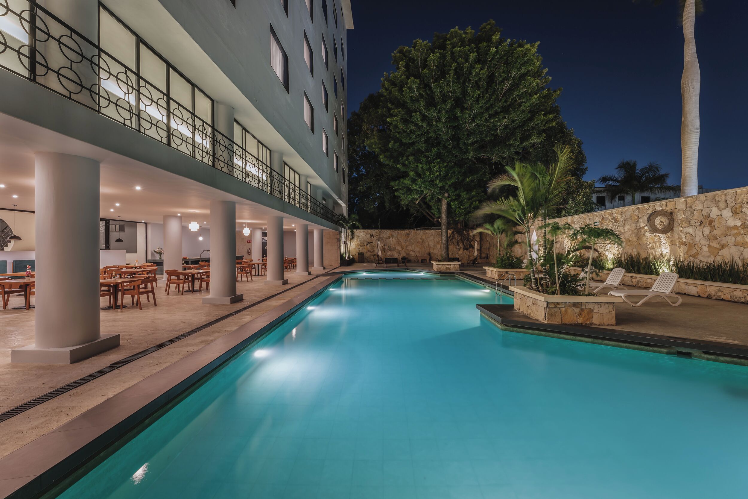 A swimming pool with a covered area, outdoor seating, and a stone wall with palm trees.