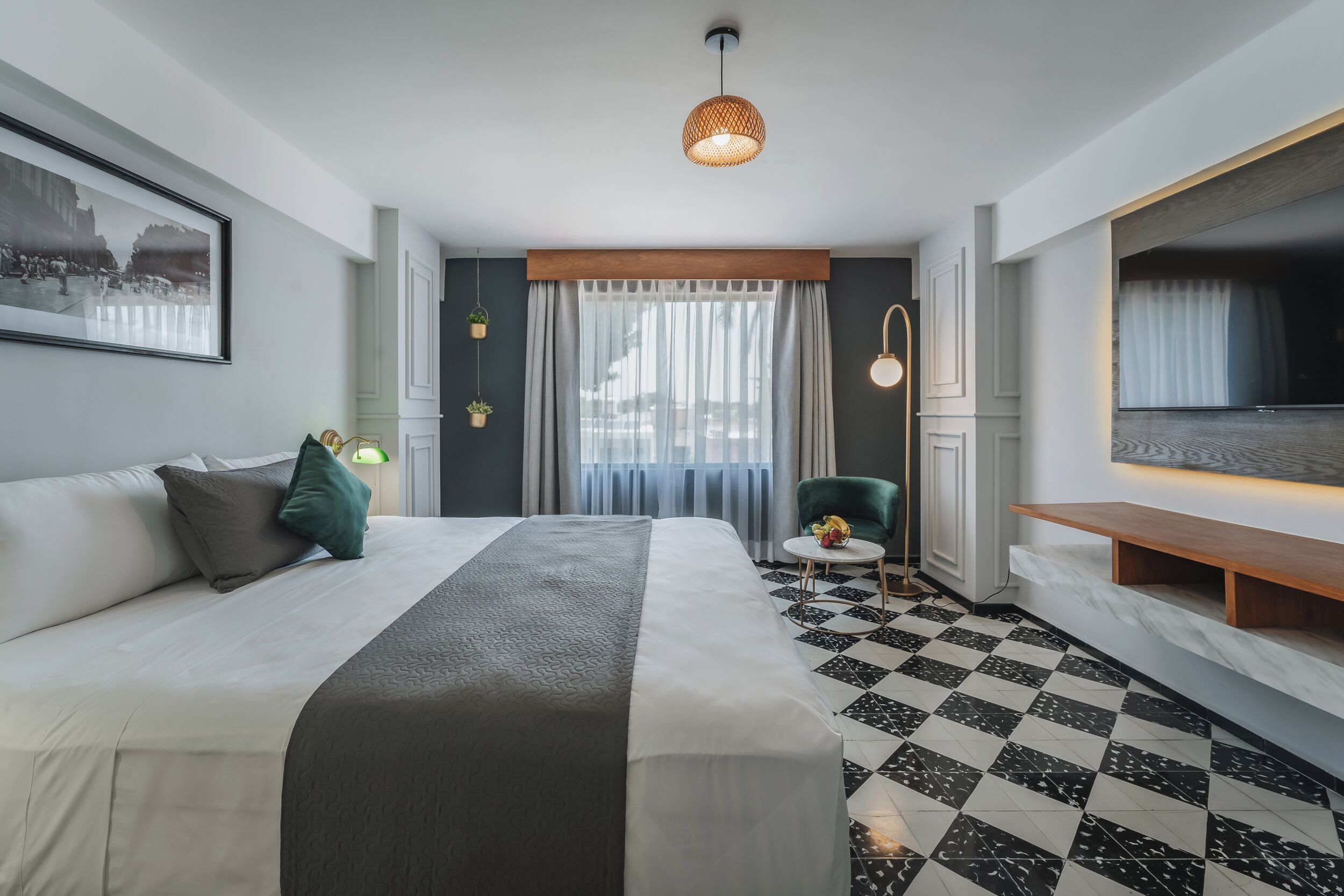 Una habitación de hotel moderna con una cama grande, una mesita redonda pequeña, una silla, un televisor y un ventanal con cortinas traslúcidas.