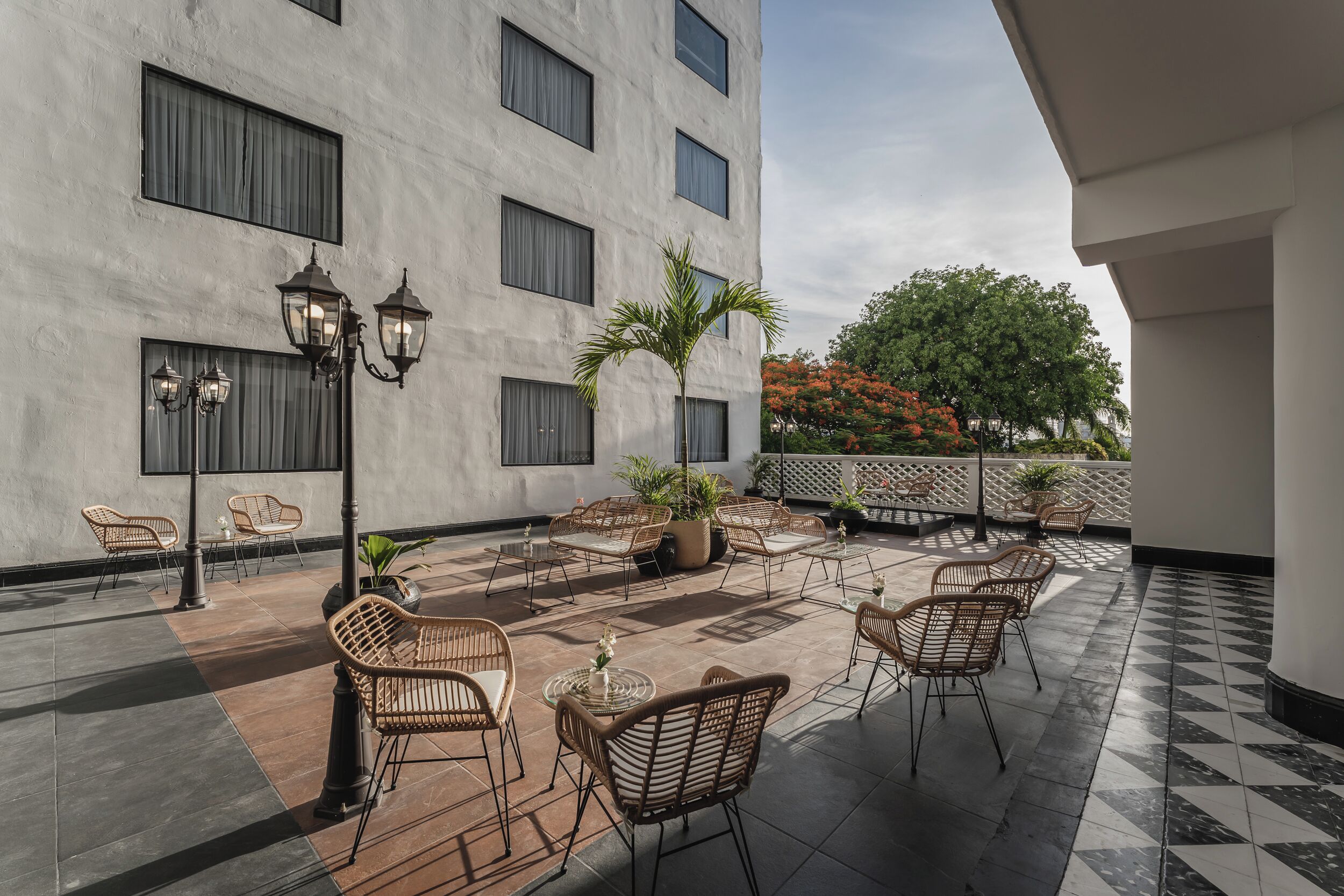 A rooftop terrace with wicker chairs, potted plants, and a view of a building with large windows and a tree with orange flowers.