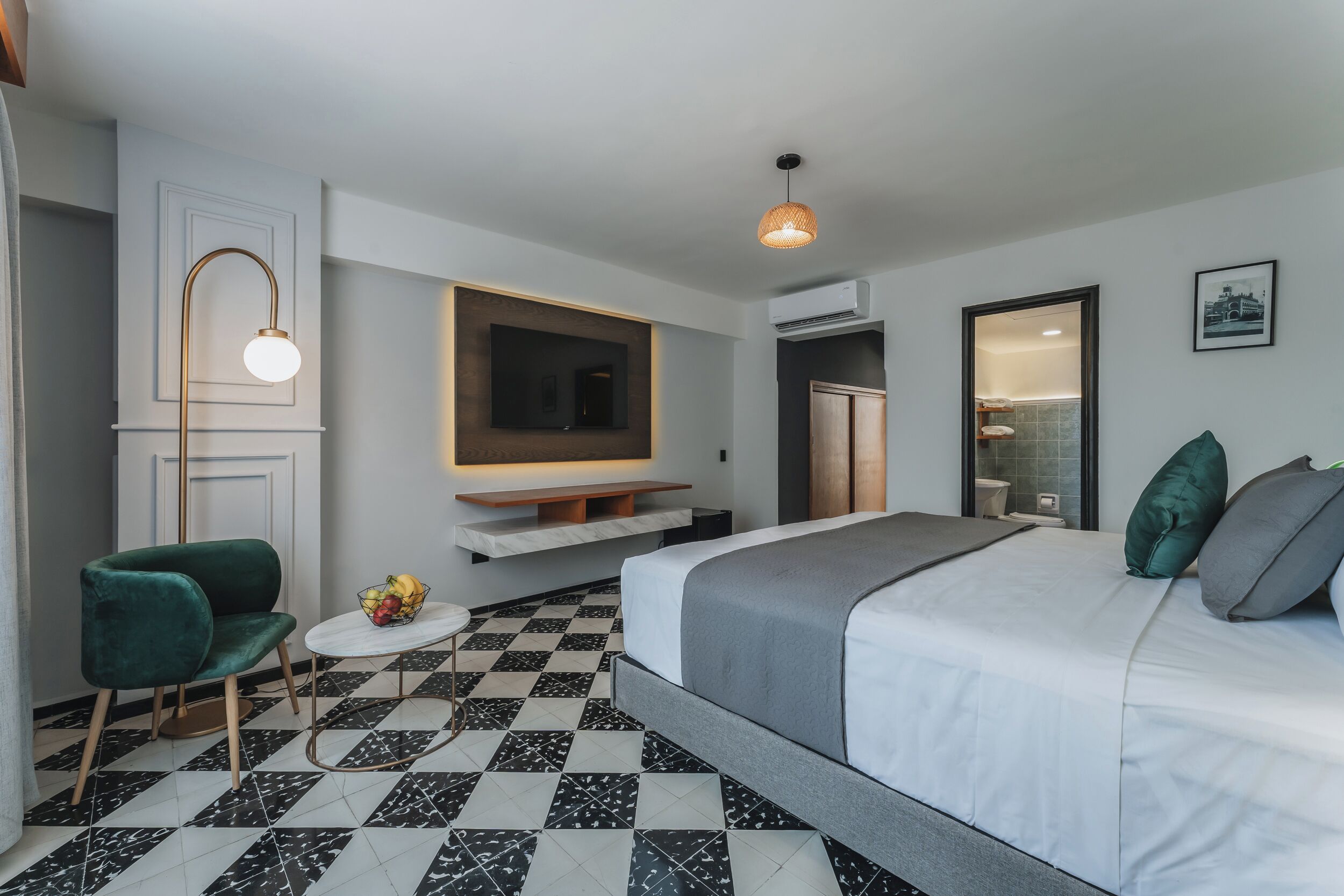 A modern hotel room with a large bed, a green armchair, a small round table with fruit, and a mounted TV.