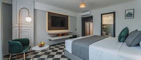 A modern hotel room with a large bed, a green armchair, a small round table with fruit, and a mounted TV.