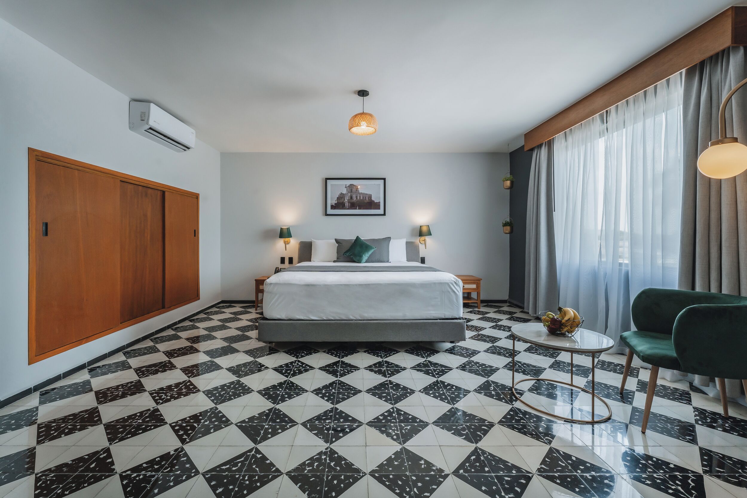 A hotel room with a checkered floor, a bed with a white headboard, a green chair, a small round table with fruit, and a large window with curtains.