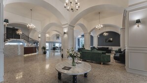 Eine geräumige Lobby mit hohen Decken, Kronleuchtern und einem Marmorboden. Es gibt einen Empfangstresen, grüne Sofas und einen runden Tisch mit Blumen.