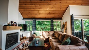 A cozy living room with a wooden ceiling, a fireplace, a large sofa, and a view of greenery outside.