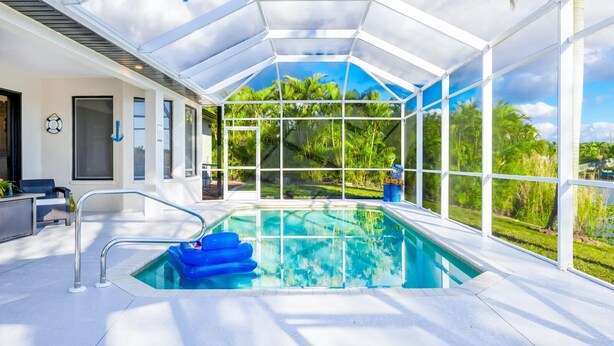 A covered swimming pool with a glass enclosure, a lounge area with outdoor furniture, and a view of lush greenery. - Canal-Front Bright Escape 3+bed/2bath -Heated Pool-Family Fun! Pet friendly! (Cape Coral)