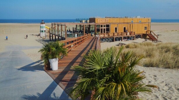 Un bâtiment situé en bord de mer, doté d’une terrasse en bois qui mène à une zone abritée, le tout entouré de sable et de végétation.