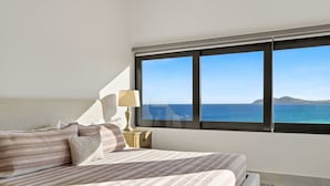 Une chambre avec une grande fenêtre, un lit, une table de chevet avec une lampe, et une vue sur la mer.