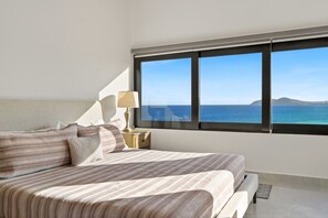 A bedroom with a large window, a bed, a bedside table with a lamp, and a view of the sea.