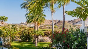 A lush garden with palm trees and flowering plants, a white column, and a view of a coastal town in the background.