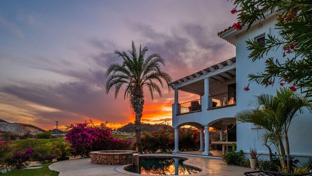 A two-story building with a balcony, surrounded by a garden with a pool and palm trees.