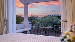 A balcony with a white sofa and a small table, overlooking a landscape with trees and hills during sunset.