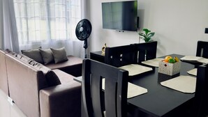 A modern living room with a sofa, a dining table with chairs, a flat-screen TV, and a standing fan.