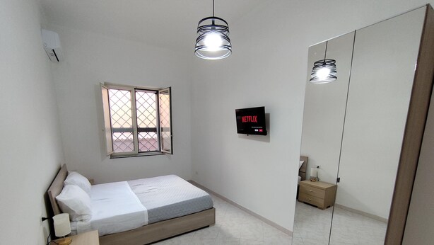 A bedroom with a bed, a bedside table, a nightstand, a window with a view, a wall-mounted TV, and a large mirror. - Vacation Home Southern Geko (Torre del Greco)