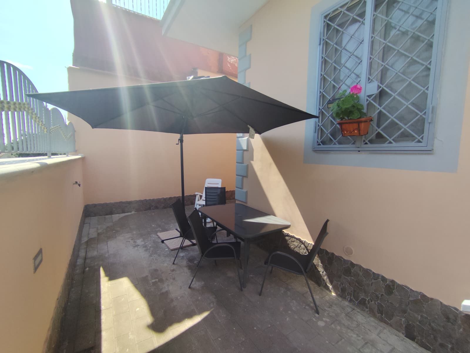 Un espace patio avec une table et des chaises, un grand parasol et une fenêtre agrémentée d’une plante en pot.