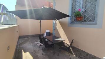 Un espace patio avec une table et des chaises, un grand parasol et une fenêtre agrémentée d’une plante en pot.