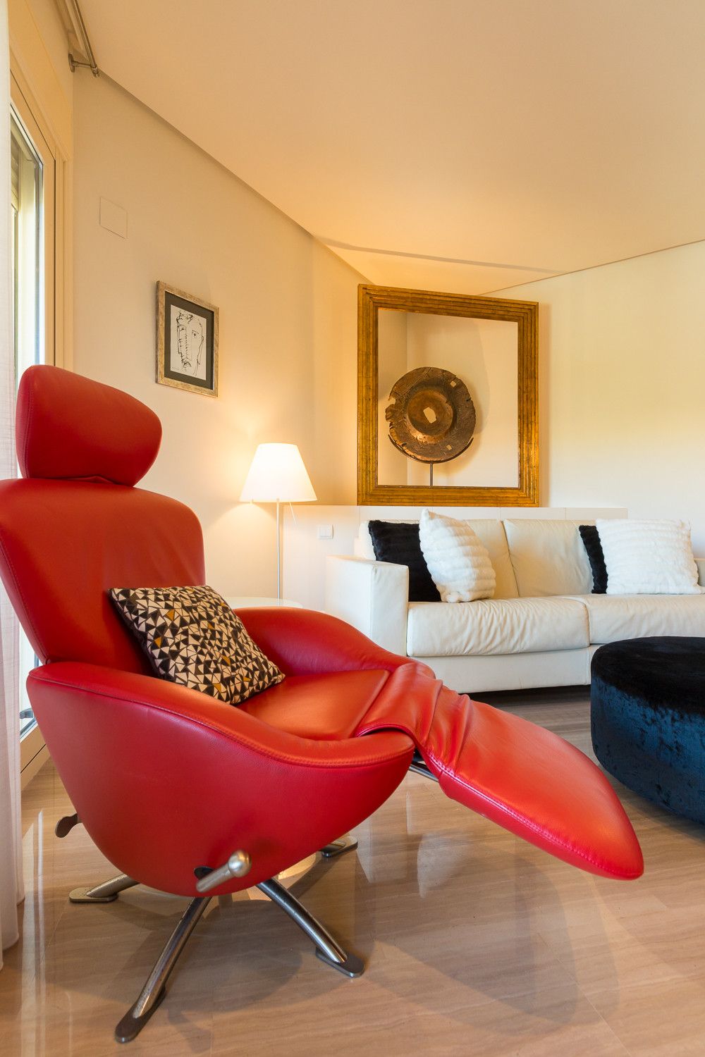 Un salon moderne avec un fauteuil rouge, un canapé blanc et un pouf bleu. On y trouve une lampe et une œuvre d’art encadrée accrochée au mur.