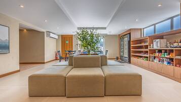A spacious living area with a large beige sectional sofa, a dining table with chairs, a kitchen area with shelves stocked with food items, and a large window providing natural light.