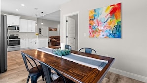 A modern kitchen with white cabinets, a wooden dining table, and a colorful abstract painting on the wall. - - August Deals - Beach Retreat with private Hot Tub & 5 min. to Navarre Beach (Navarre)
