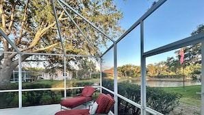 A covered outdoor area with two red-striped lounge chairs and a small table, overlooking a body of water and a grassy area with trees.