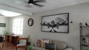 A living room with a ceiling fan, a large framed picture of an airplane, a clock, and a bookshelf with a bird sculpture.