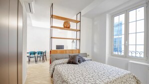 A bedroom with a bed, a desk, and a chair. There is a window with a view of buildings.