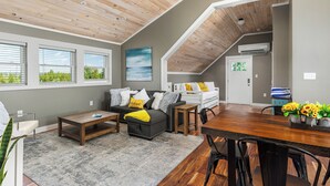 A cozy loft-style living room with a wooden ceiling, a gray sofa with yellow and white cushions, a wooden coffee table, and a dining table with chairs. - Pleasant Valley Resort (Arcadia)