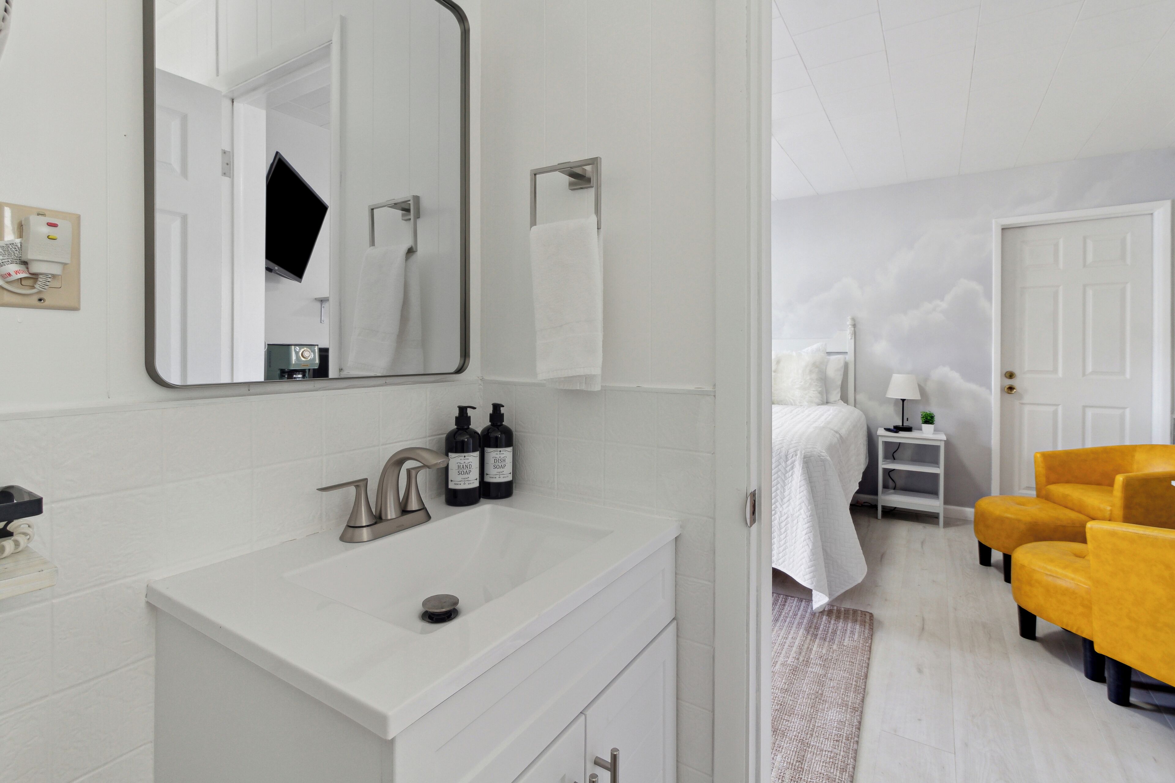 Une salle de bain moderne avec un lavabo blanc, un miroir et un porte-serviettes. On y trouve un lit recouvert d’une couverture blanche et une table de chevet avec une lampe.