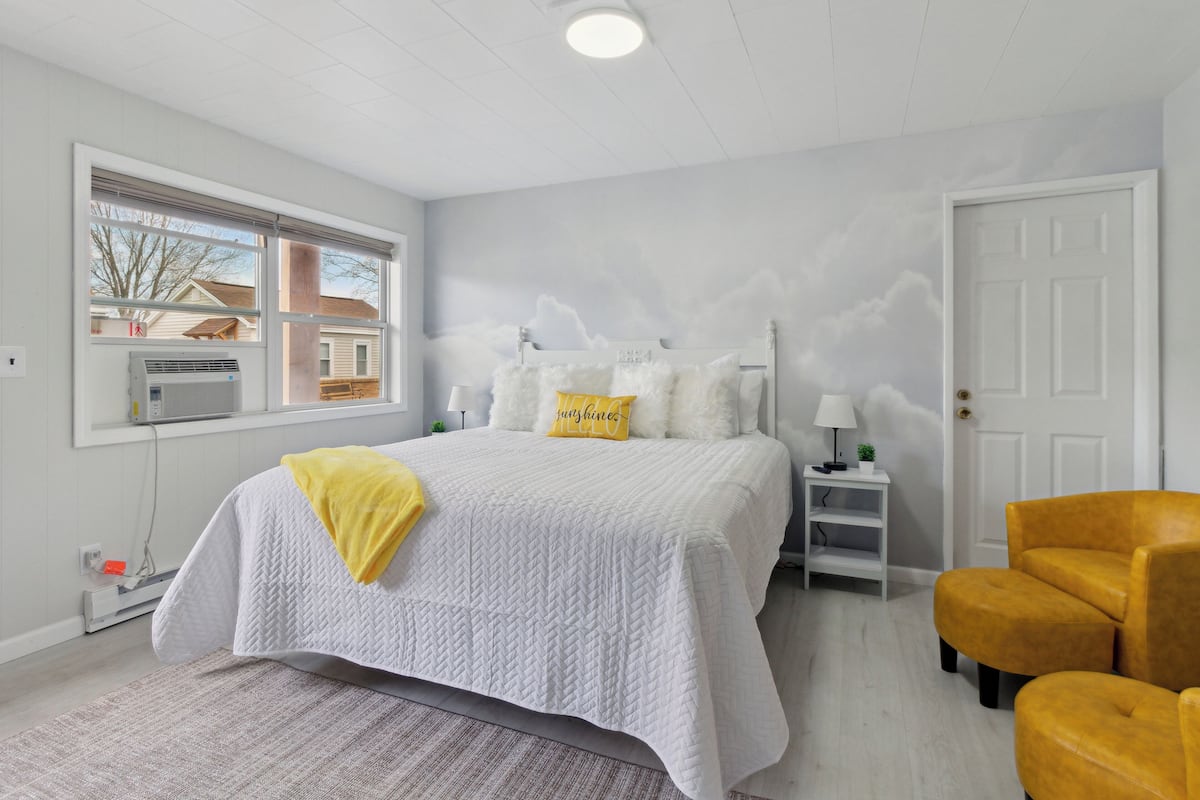 A bedroom with a large bed, a window with blinds, a white door, and a yellow armchair.
