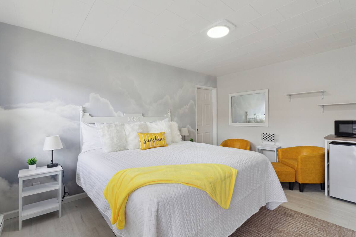 A modern hotel room with a large bed, a bedside table with a lamp, a small plant, and a yellow throw blanket.
