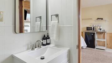 Un baño moderno con un lavamanos blanco, un espejo y un perchero. Hay un microondas y un armario pequeño en la habitación contigua.