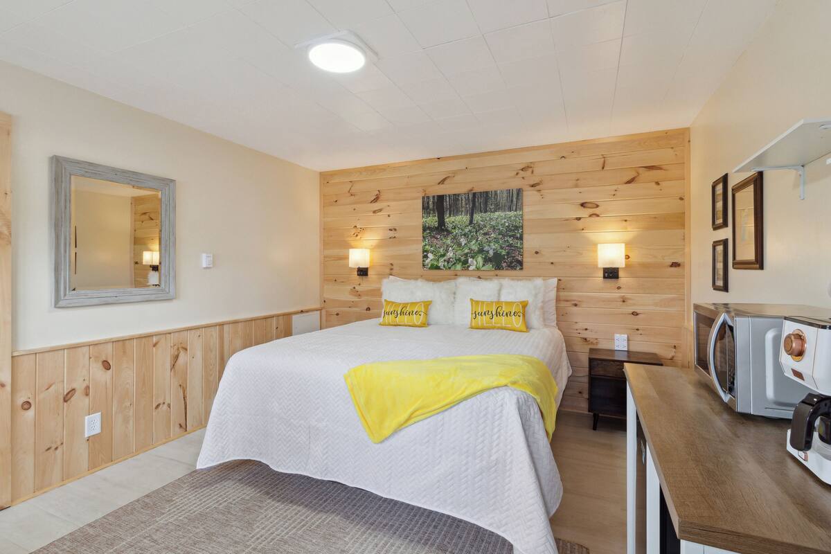 A bedroom with a wooden wall panel, a bed with white bedding and two pillows, a bedside table with a lamp, and a microwave on a wooden countertop.