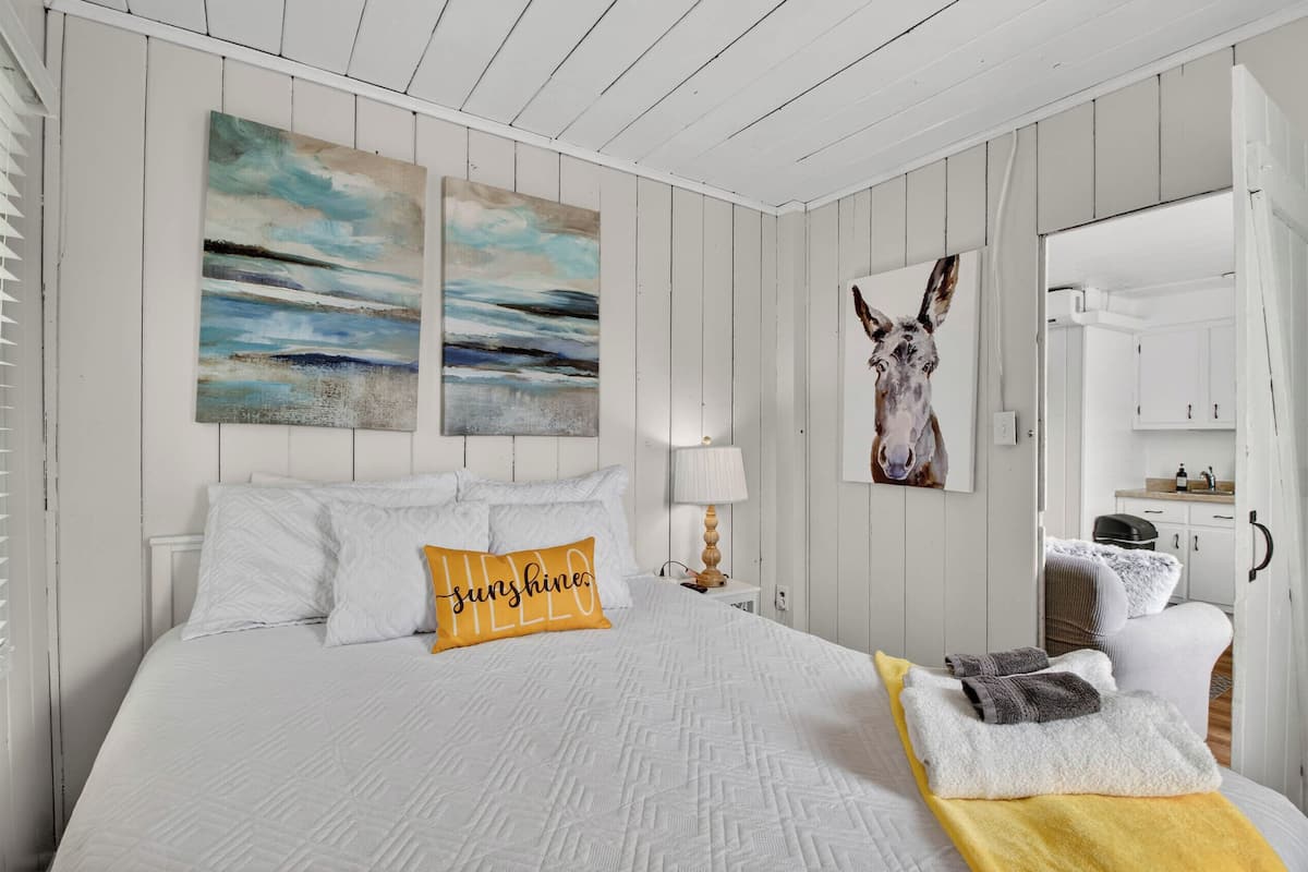A bedroom with a bed, a nightstand, a lamp, a painting, and a bathroom visible through an open door.