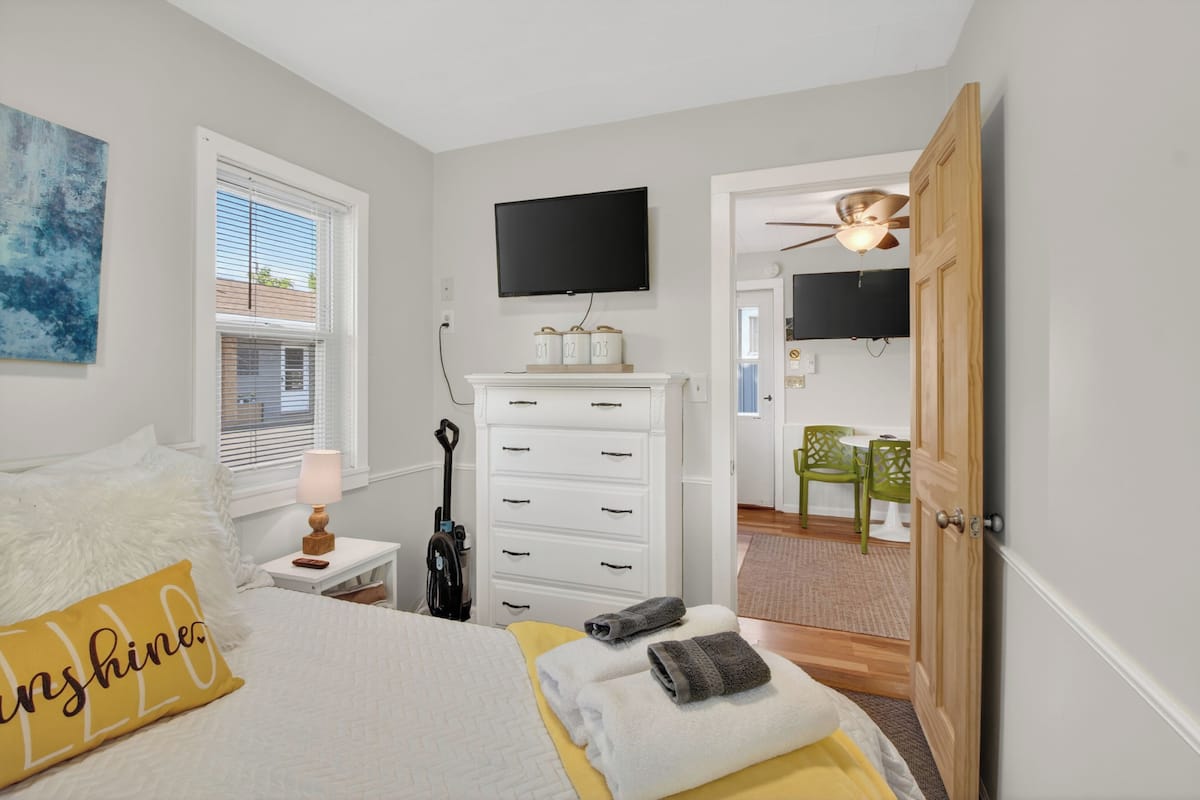 A bedroom with a bed, a TV, a dresser, and a door leading to a dining area.