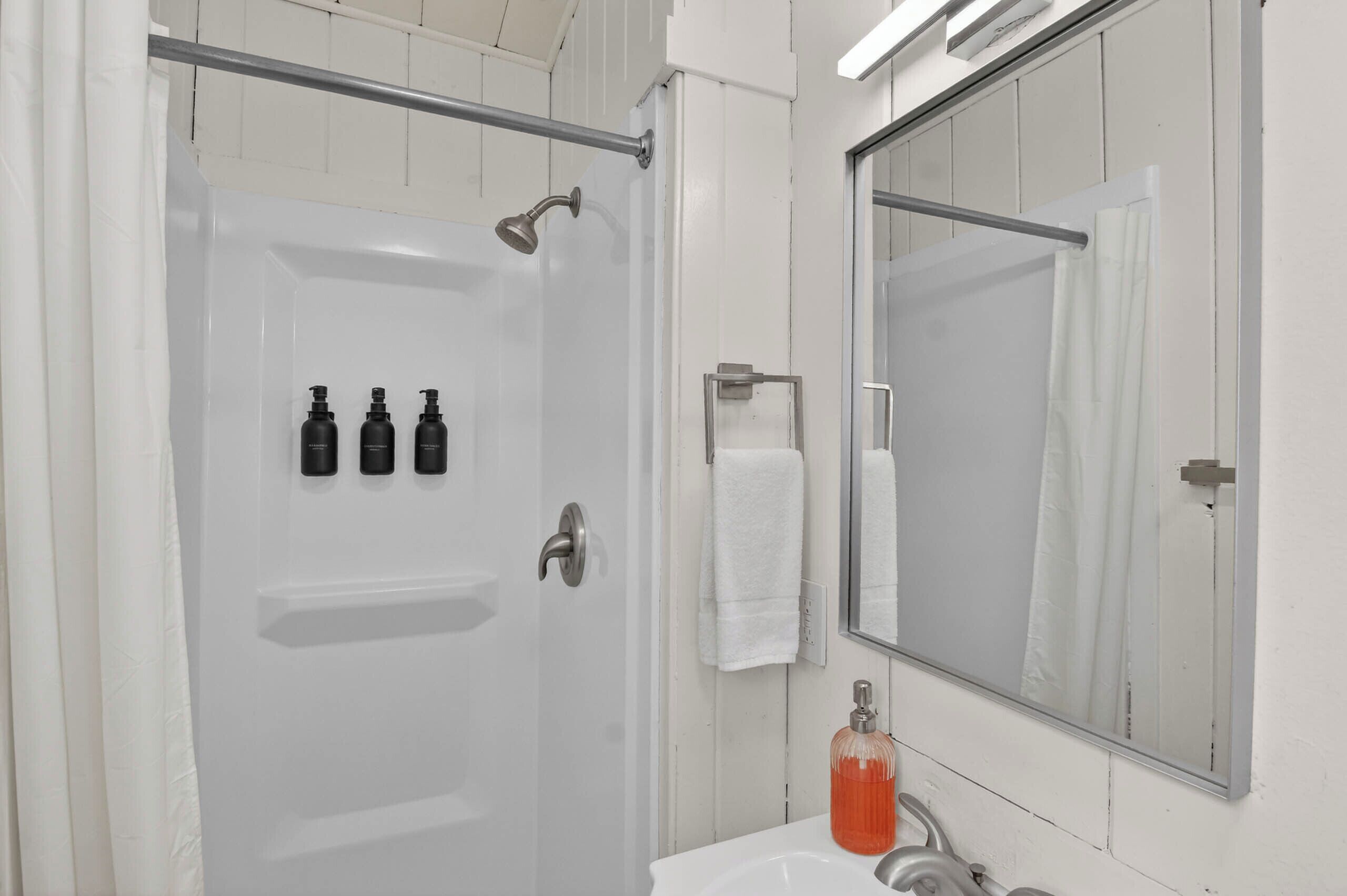 Un baño con ducha, lavamanos, espejo y tres botellas en la pared.