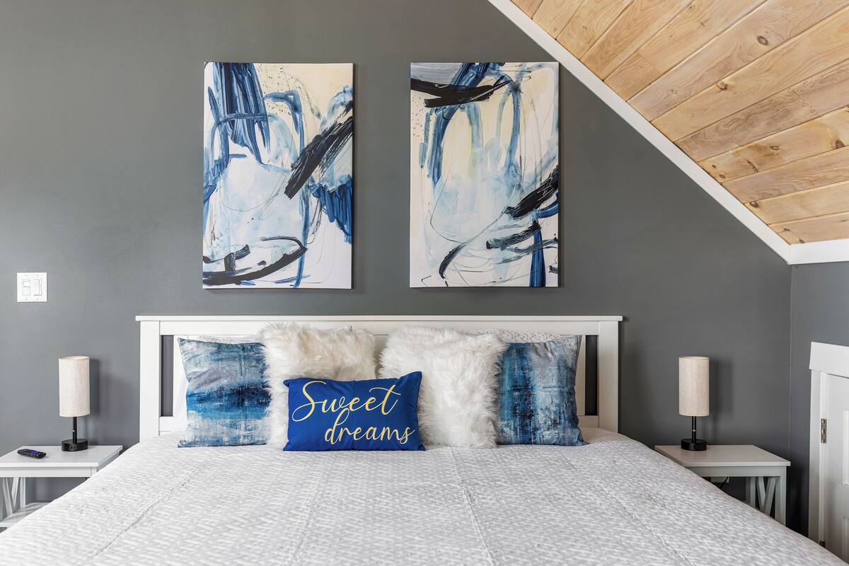 A bedroom with a bed, two wall-mounted paintings, a nightstand, a lamp, and a pillow with the text "Sweet dreams".