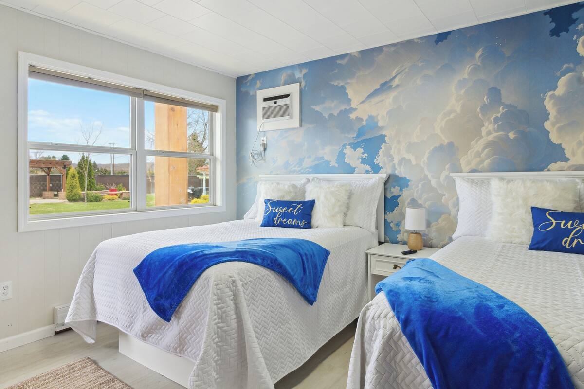 A bedroom with two beds, a large window, and a mural of clouds on the wall.