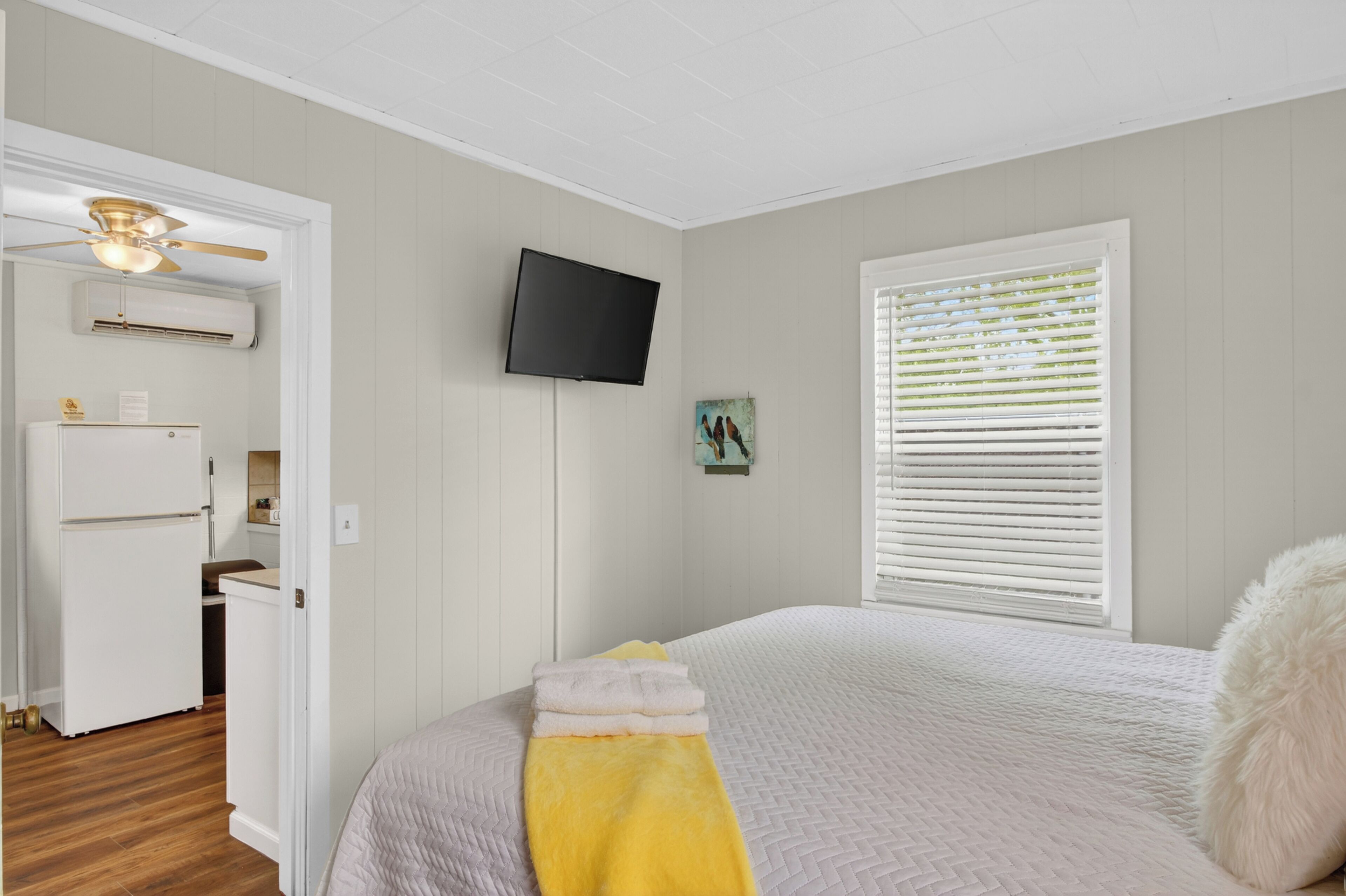 A bedroom with a bed, a television mounted on the wall, a window with blinds, and a kitchen area visible through an open door.