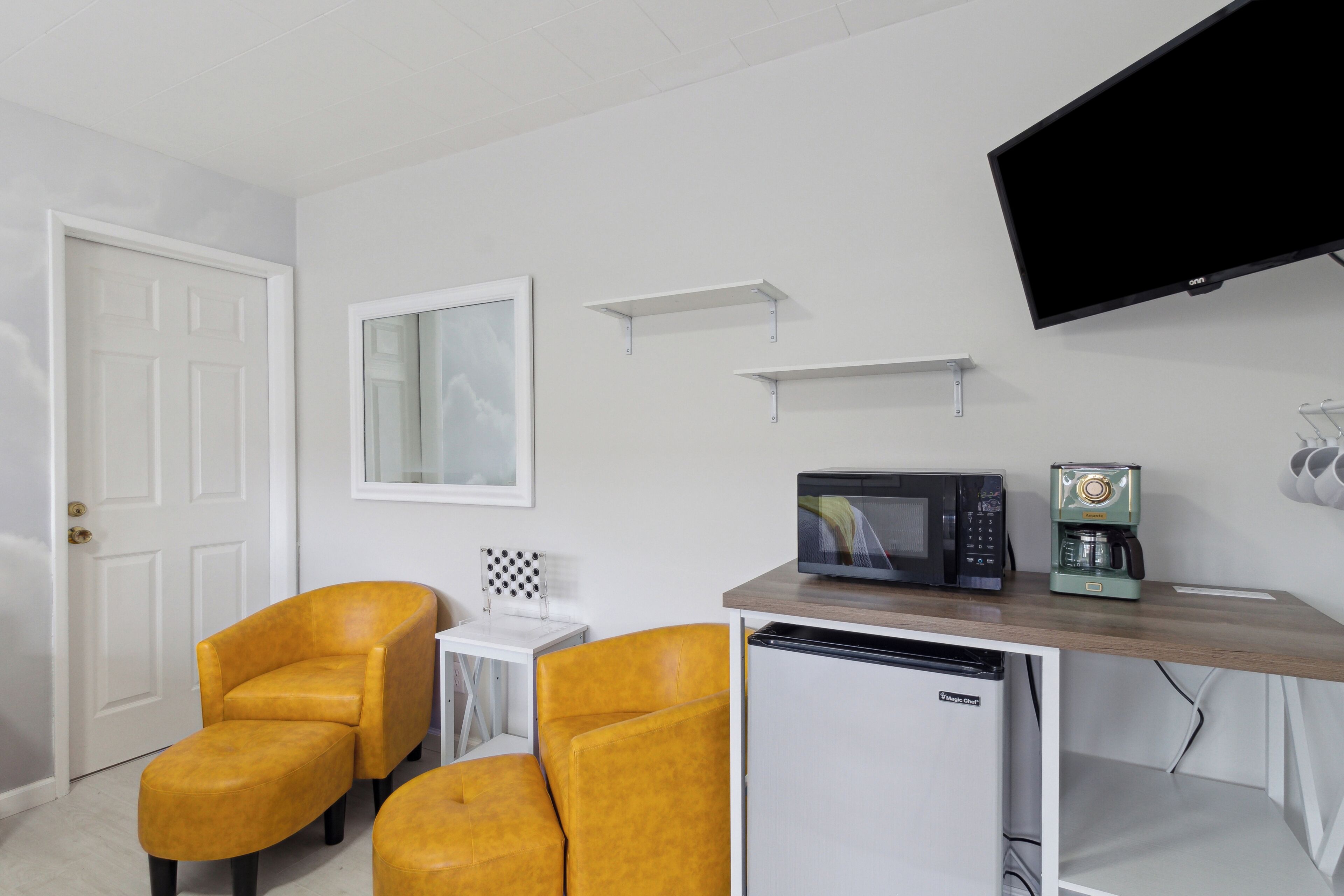 A compact room with a white door, two yellow armchairs, a small white table, a microwave, a coffee machine, and a flat-screen TV mounted on the wall.