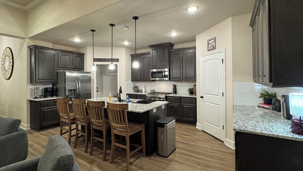 A modern kitchen with dark cabinets, stainless steel appliances, and a granite countertop. There are wooden chairs around a central island, and a trash can is visible.
